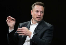 Man in suit speaking, black background, gesturing with hands.