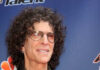 A man with curly hair and glasses smiling at a red carpet event