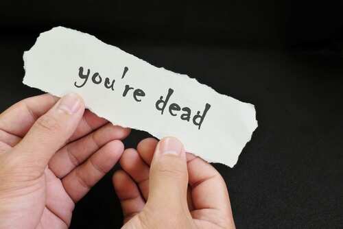 shutterstock_2214861359.jpg A person holding a torn piece of paper with the words 'you're dead' written on it
