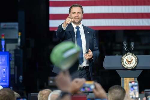 A speaker gestures towards the audience during a political event with an American flag in the background