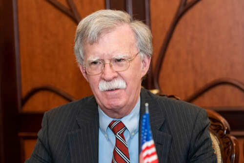 A man in a suit with glasses sitting at a table with an American flag