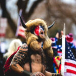 Individual in a fur hat and face paint holding an American flag at a protest