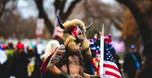 shutterstock_1888591864 (1).jpg Individual in a fur hat and face paint holding an American flag at a protest