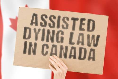 A hand holding a cardboard sign that reads 'Assisted Dying Law in Canada' with a blurred Canadian flag in the background