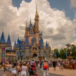 Another Guest Dies At Disney… Crowd of visitors in front of a large Disney castle under a cloudy sky