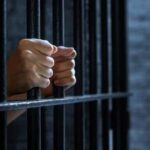 Hands gripping prison bars in a dark setting
