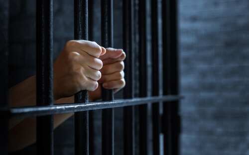 shutterstock_2551401195.jpg Hands gripping prison bars in a dark setting