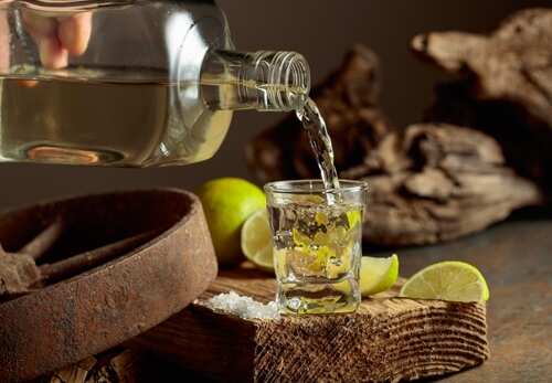 A shot glass being filled with tequila next to lime slices and salt