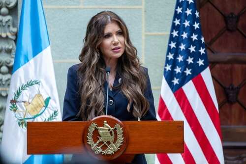 shutterstock_2646539471.jpg A woman speaking at a podium with national flags in the background
