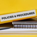 A yellow binder labeled 'POLICIES & PROCEDURES' next to notebooks and a pen