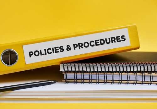 shutterstock_2424424421.jpg A yellow binder labeled 'POLICIES & PROCEDURES' next to notebooks and a pen