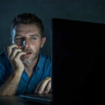 Man sitting in front of a laptop with a focused expression in a dimly lit room