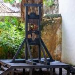 A wooden guillotine displayed in an outdoor area surrounded by greenery