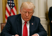 Man in suit signing document at desk with flag