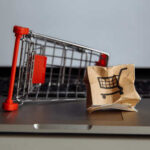 A miniature shopping cart next to a crumpled paper bag with a shopping cart icon