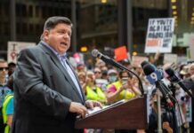 A speaker addressing a crowd at a political rally with protest signs in the background