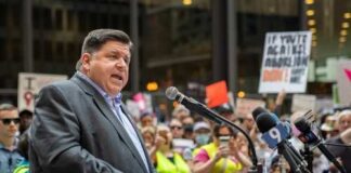 A speaker addressing a crowd at a political rally with protest signs in the background