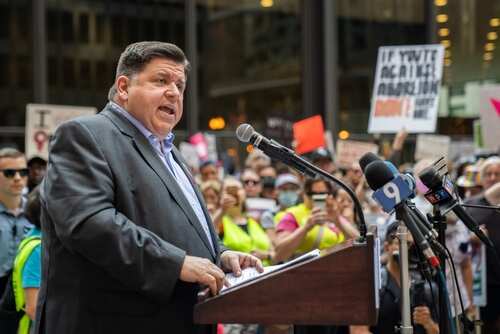 A speaker addressing a crowd at a political rally with protest signs in the background