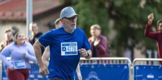 An older man running in a marathon wearing a blue shirt and a race bib