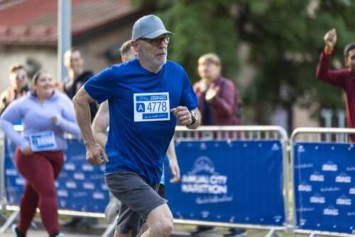 An older man running in a marathon wearing a blue shirt and a race bib
