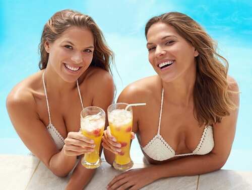 Two women smiling and holding tropical drinks by the pool