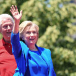 Hillary Clinton waving at an outdoor event with Bill Clinton in the background