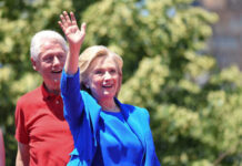 Hillary Clinton waving at an outdoor event with Bill Clinton in the background