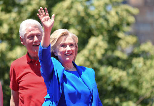 shutterstock_287370860.jpg Hillary Clinton waving at an outdoor event with Bill Clinton in the background