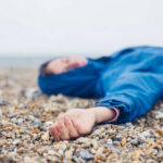 A person in a blue jacket lying on a pebbled beach with a blurred background