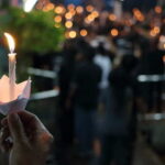 A hand holding a lit candle in a paper holder at a vigil