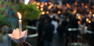 A hand holding a lit candle in a paper holder at a vigil