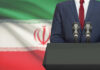 A suited individual standing behind a podium with microphones in front of the Iranian flag