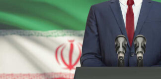 A suited individual standing behind a podium with microphones in front of the Iranian flag