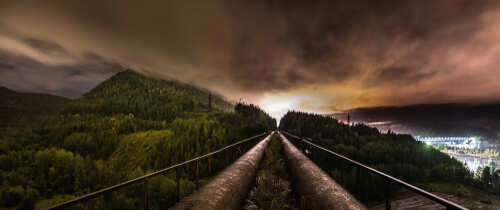 Panoramic view of industrial pipes leading towards a mountainous landscape under a cloudy night sky