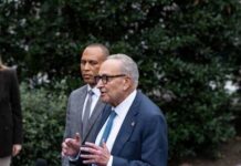 Two men speaking at an outdoor press conference surrounded by greenery