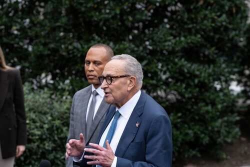 Two men speaking at an outdoor press conference surrounded by greenery