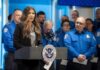 A woman speaking at a podium with law enforcement officers in the background