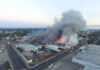 A large industrial fire with thick smoke rising into the sky and firefighters responding